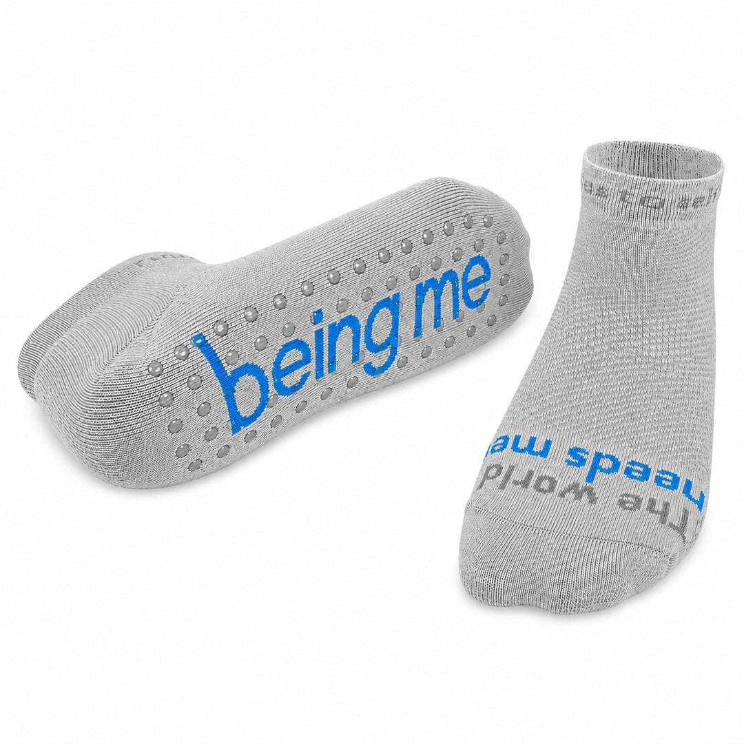 A pair of light gray ankle socks with textured grip dots on the sole and blue text reading 'being me' and 'The world needs me', shown on a plain white background.